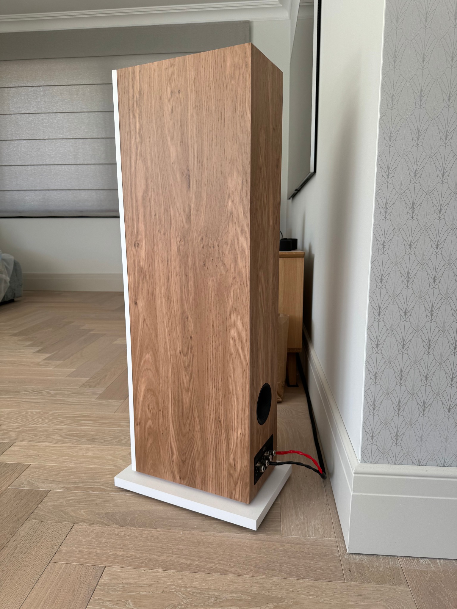 Tył drewnianej kolumny głośnikowej z podłączonymi kablami, ustawionej na podłodze w jodełkę, z widocznym fragmentem ściany z wzorzystą tapetą.