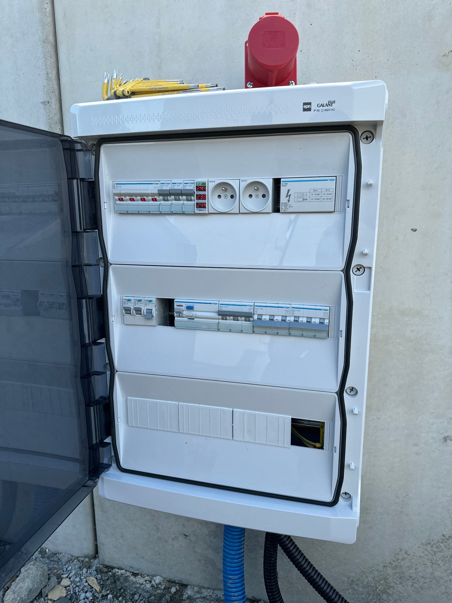 Otwarta skrzynka elektryczna z widocznymi bezpiecznikami, gniazdkami i przewodami, zamontowana na szarej ścianie, z narzędziami leżącymi na górze i niebieskim peszlem doprowadzonym od dołu.