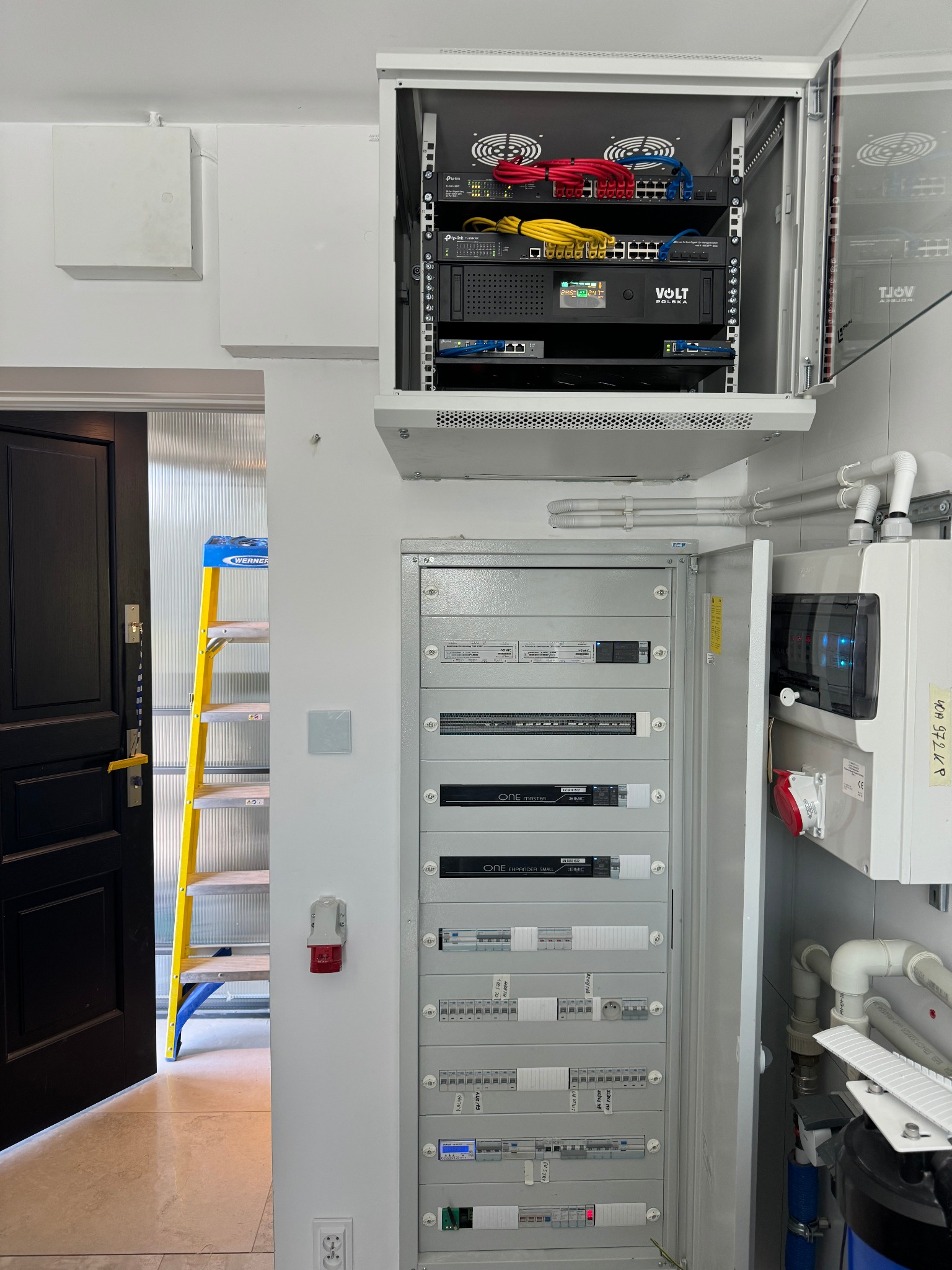Otwarta szafa rack z urządzeniami sieciowymi, w tym routerami i przełącznikami, nad rozdzielnicą elektryczną z licznymi bezpiecznikami i modułami, obok drabina i fragment drzwi wejściowych.