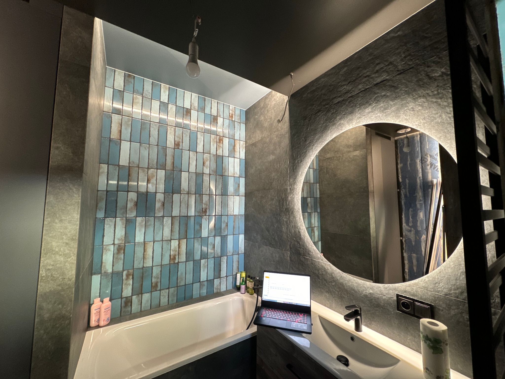 Nowoczesna łazienka z niebieskimi płytkami, okrągłym lustrem z podświetleniem LED i laptopem na blacie umywalki, wisząca żarówka bez klosza na suficie.