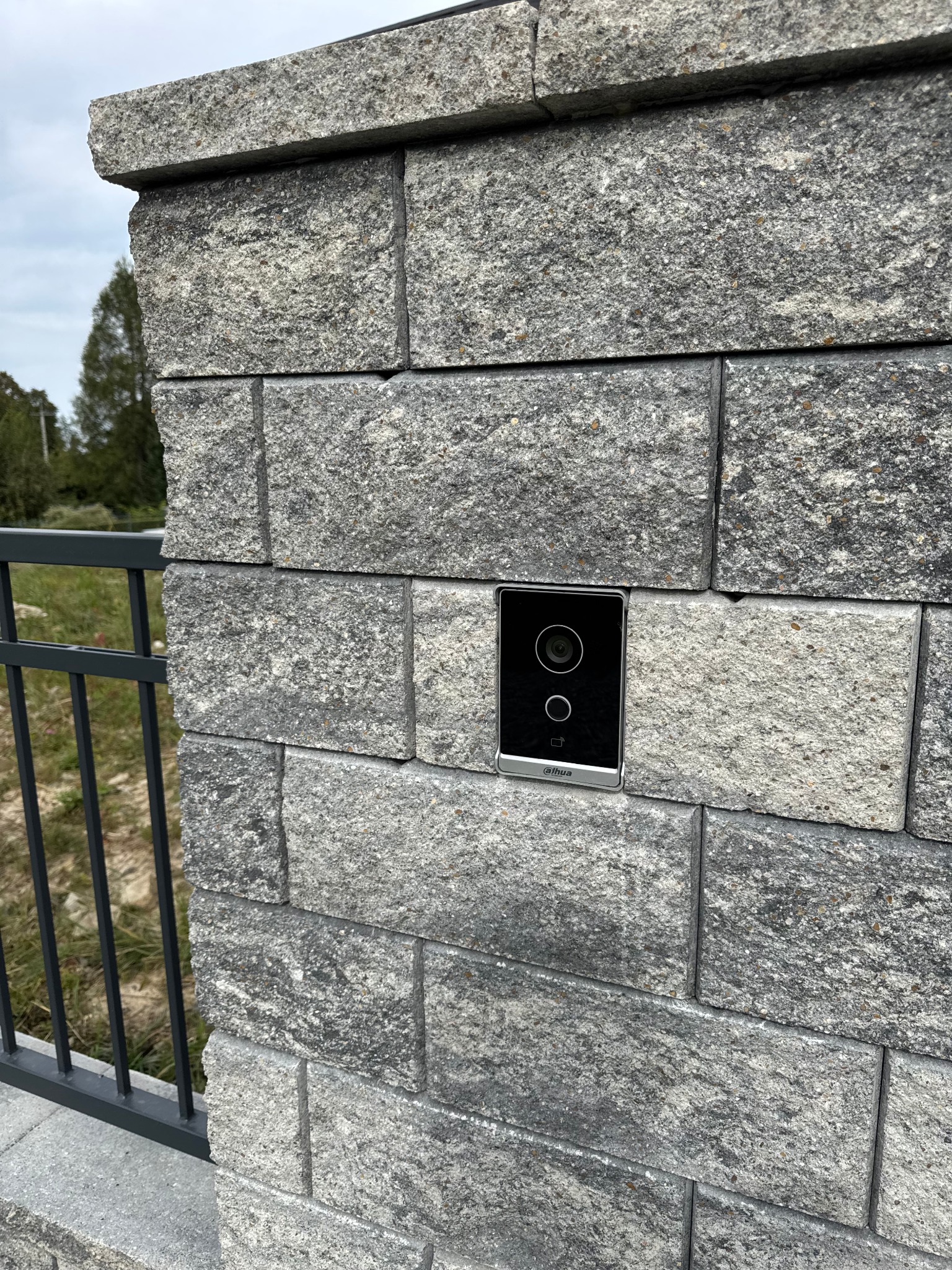 Wideodomofon Dahua zamontowany na murowanym słupku ogrodzeniowym z szarego kamienia łupanego, widok z bliska, z fragmentem czarnego ogrodzenia w tle.