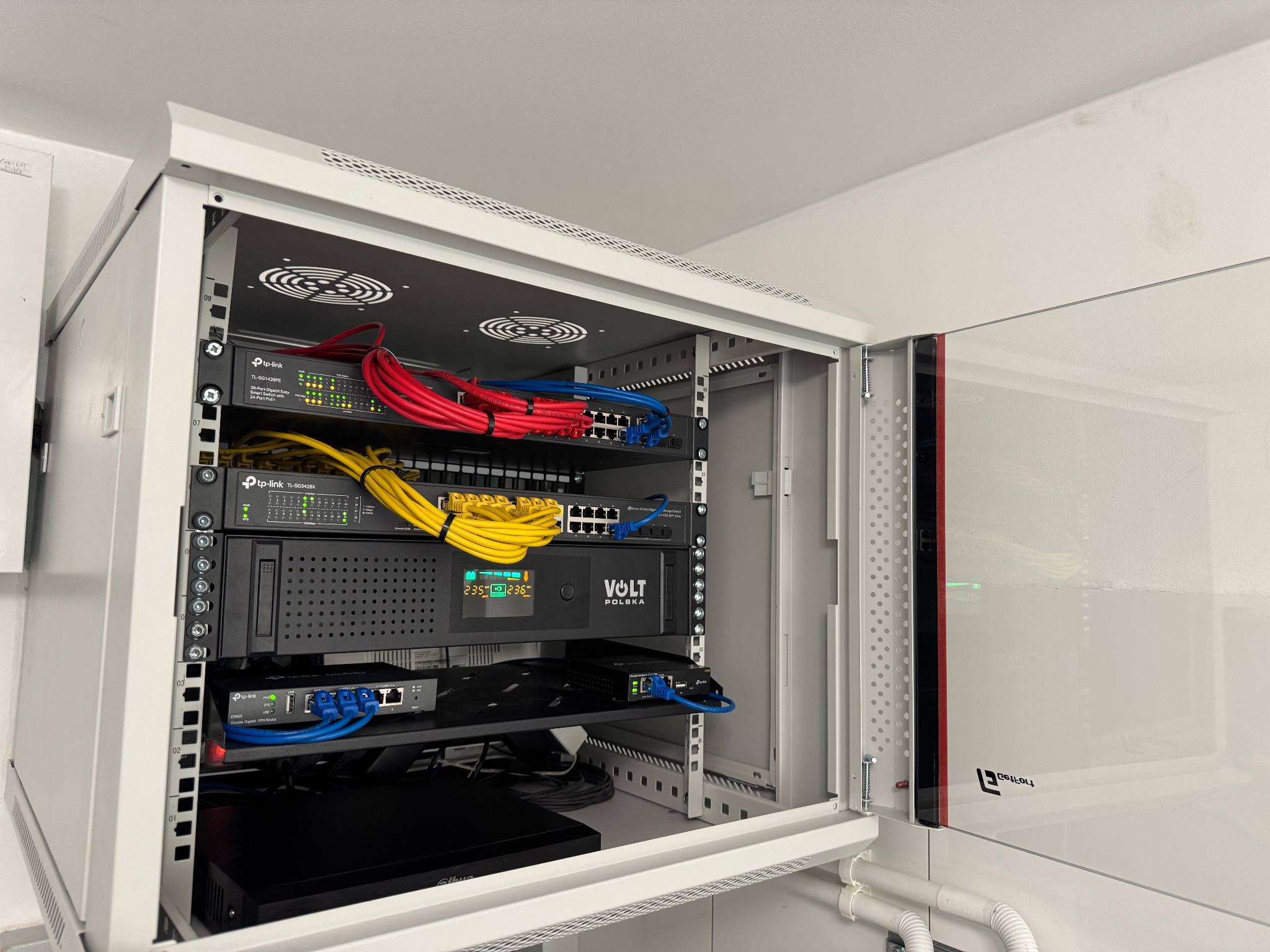 Otwarta szafa rack z zainstalowanymi urządzeniami sieciowymi, w tym switche TP-Link z kolorowymi kablami Ethernet (czerwone, niebieskie, żółte), zasilacz awaryjny Volt Polska z wyświetlaczem...