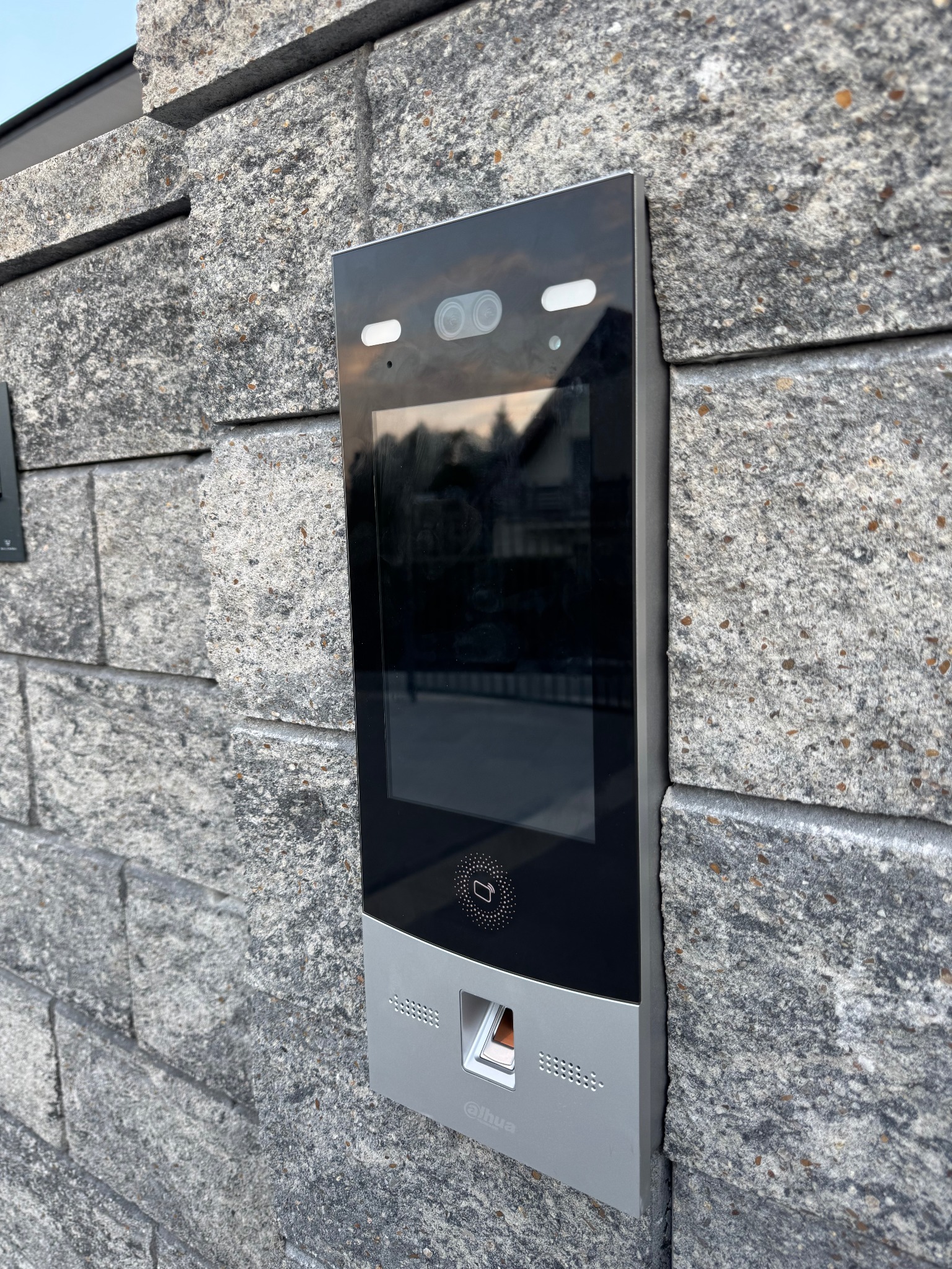 Zewnętrzny panel wideodomofonu z czytnikiem linii papilarnych i rozpoznawaniem twarzy, zamontowany na szarej, kamiennej ścianie budynku.