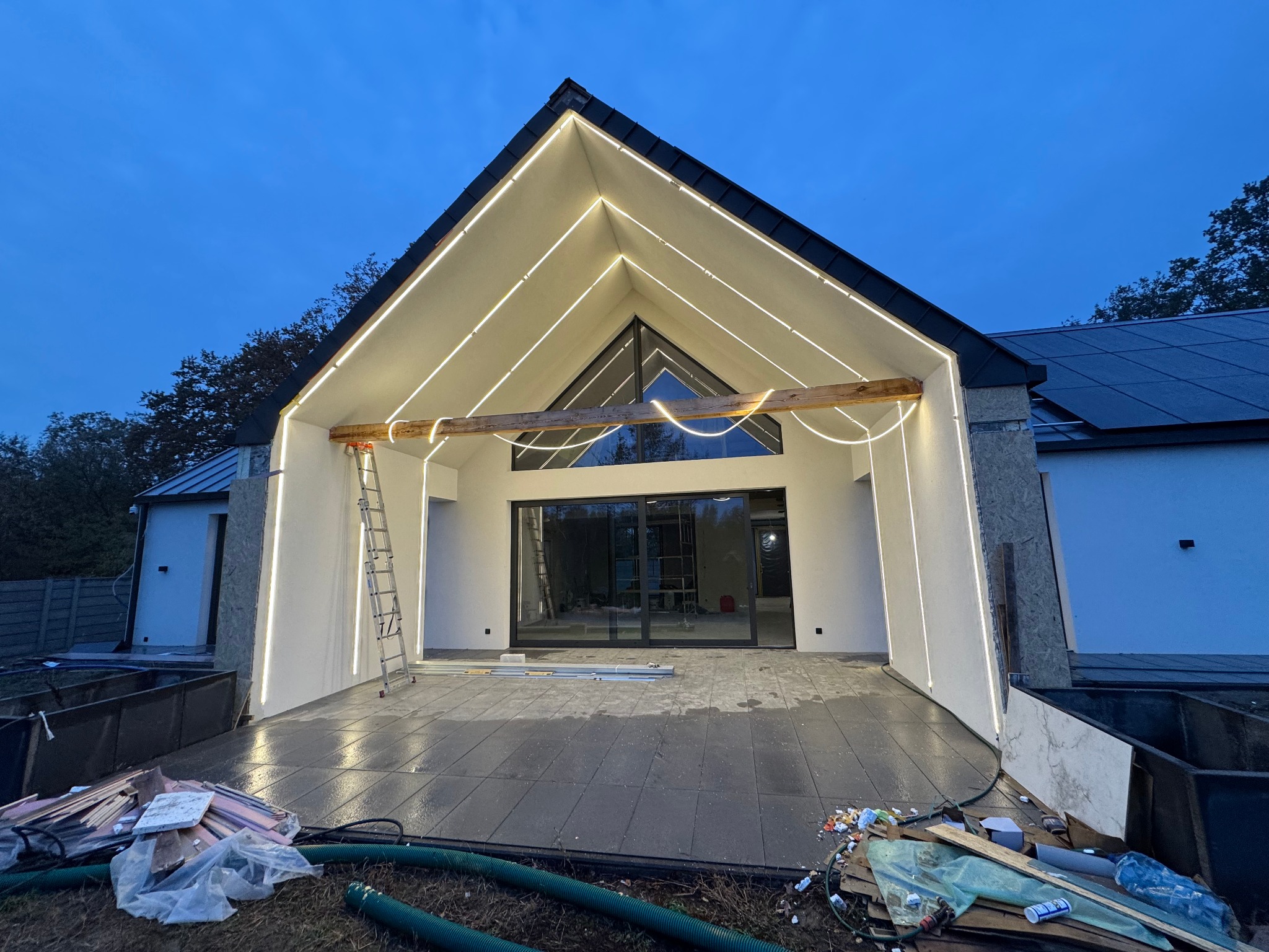 Nowoczesny dom w trakcie wykończenia z podświetleniem LED wzdłuż dachu, widoczny bałagan budowlany na tarasie i drabina oparta o ścianę.