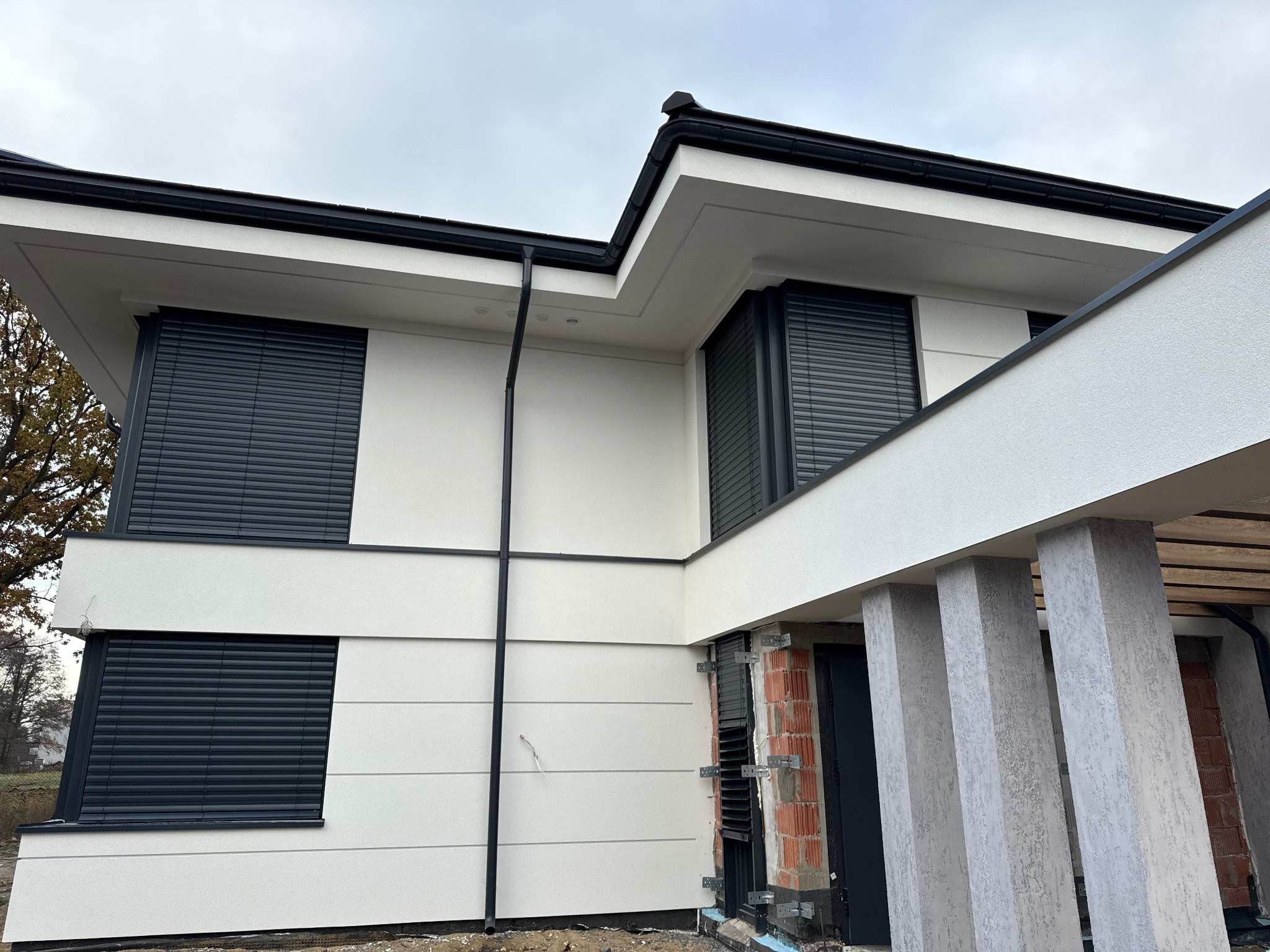 Nowoczesny dom jednorodzinny w trakcie wykończenia z zamontowanymi roletami zewnętrznymi w kolorze antracytowym i elewacją w odcieniach bieli.