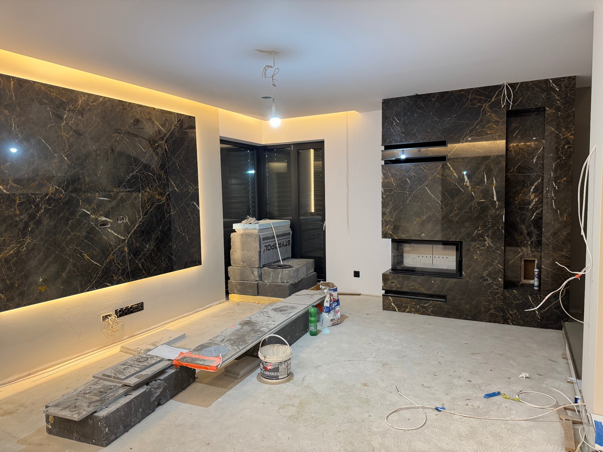 Wnętrze w trakcie remontu z panelami imitującymi czarny marmur z oświetleniem LED, widoczne kable elektryczne, materiały budowlane i kominek.