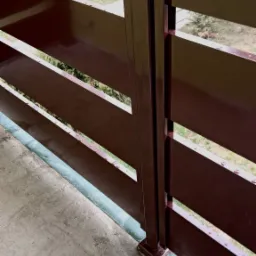 Fragment metalowej balustrady z poziomymi szczeblami, pomalowanej na brązowo, widoczny fragment betonowego podłoża i zieleń w tle.