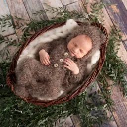 Śpiący noworodek w brązowym, futerkowym kombinezonie z uszkami, leżący w wiklinowym koszu wyłożonym białym futerkiem, otoczonym zielonymi gałązkami na drewnianym tle.