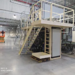 Wnętrze hali produkcyjnej z konstrukcją stalową z platformą i schodami, w tle podnośnik koszowy z pracownikiem, widoczne instalacje i elementy linii produkcyjnej.