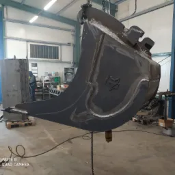 Ciężka, metalowa łyżka koparki zawieszona na łańcuchu w hali warsztatowej, widoczne ślady spawania i logo firmy.