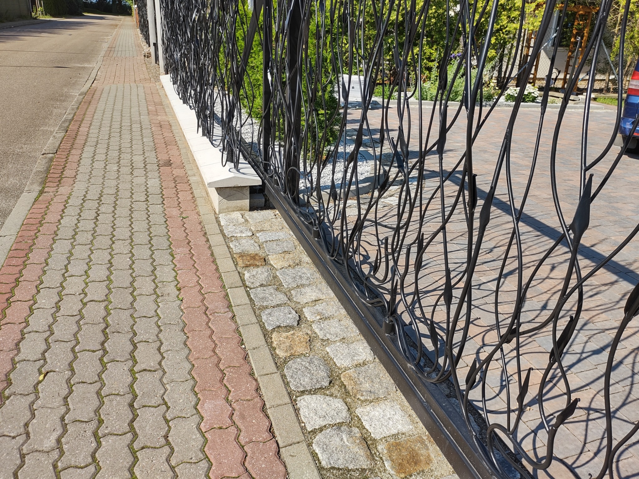 Nowoczesne, czarne ogrodzenie z metalu o nieregularnym wzorze, oddzielające chodnik z kostki brukowej od posesji z wyłożonym kamieniem podjazdem i zielenią w tle.