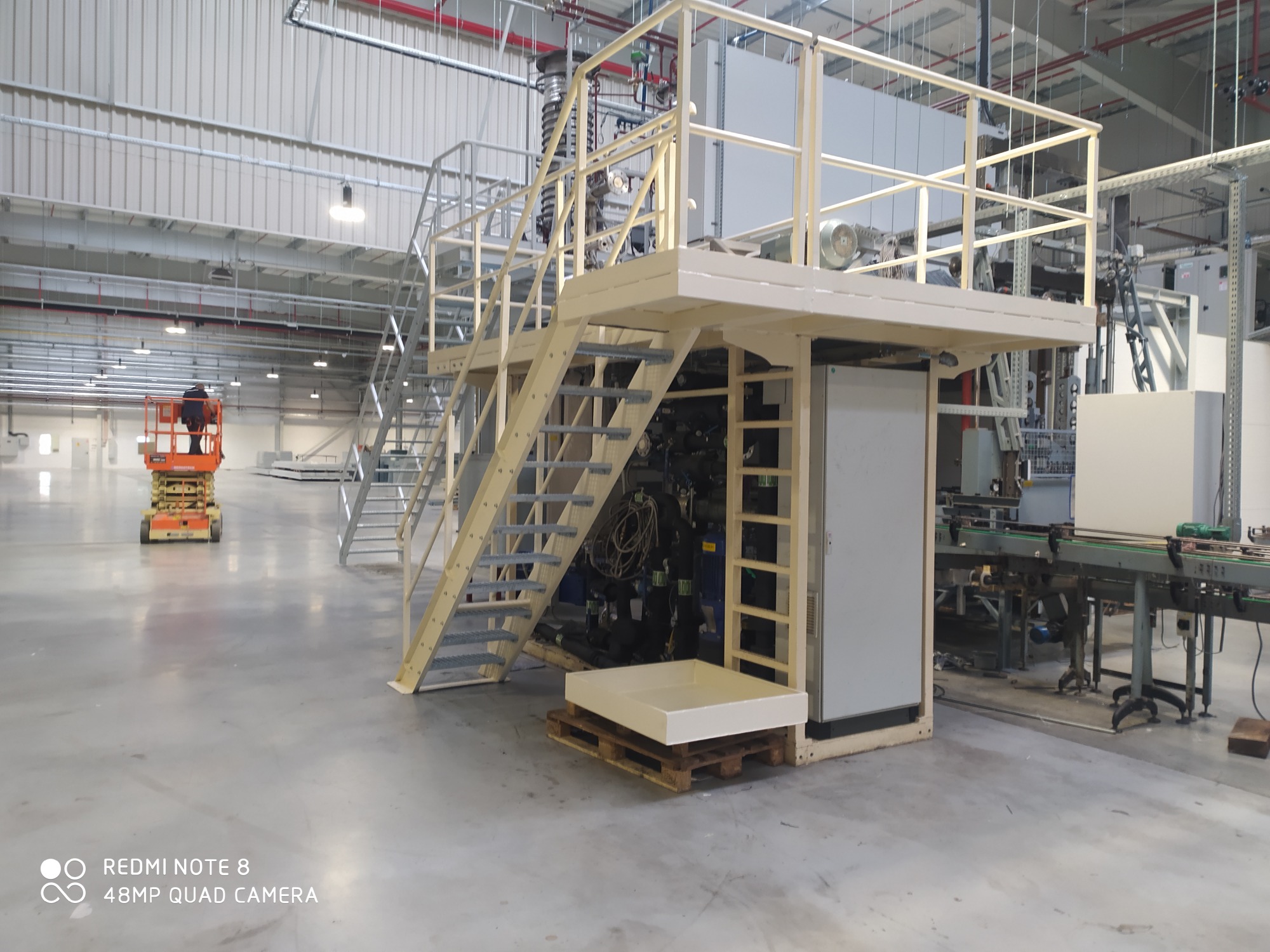 Wnętrze hali produkcyjnej z konstrukcją stalową z platformą i schodami, w tle podnośnik koszowy z pracownikiem, widoczne instalacje i elementy linii produkcyjnej.