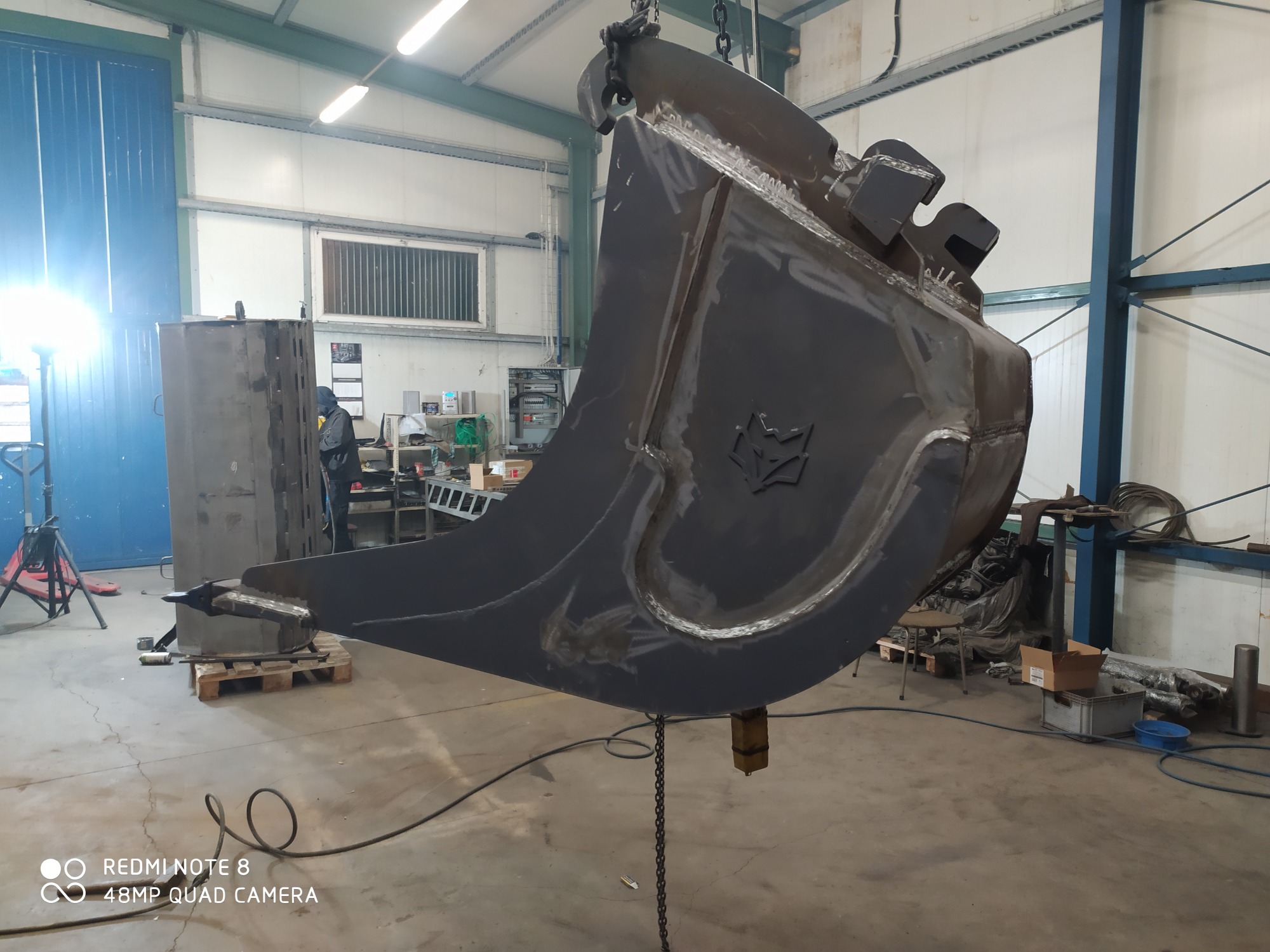 Ciężka, metalowa łyżka koparki zawieszona na łańcuchu w hali warsztatowej, widoczne ślady spawania i logo firmy.