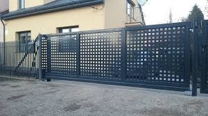 Nowoczesna, przesuwna brama wjazdowa z ażurowym wypełnieniem w kolorze grafitowym na tle jasnego budynku mieszkalnego.