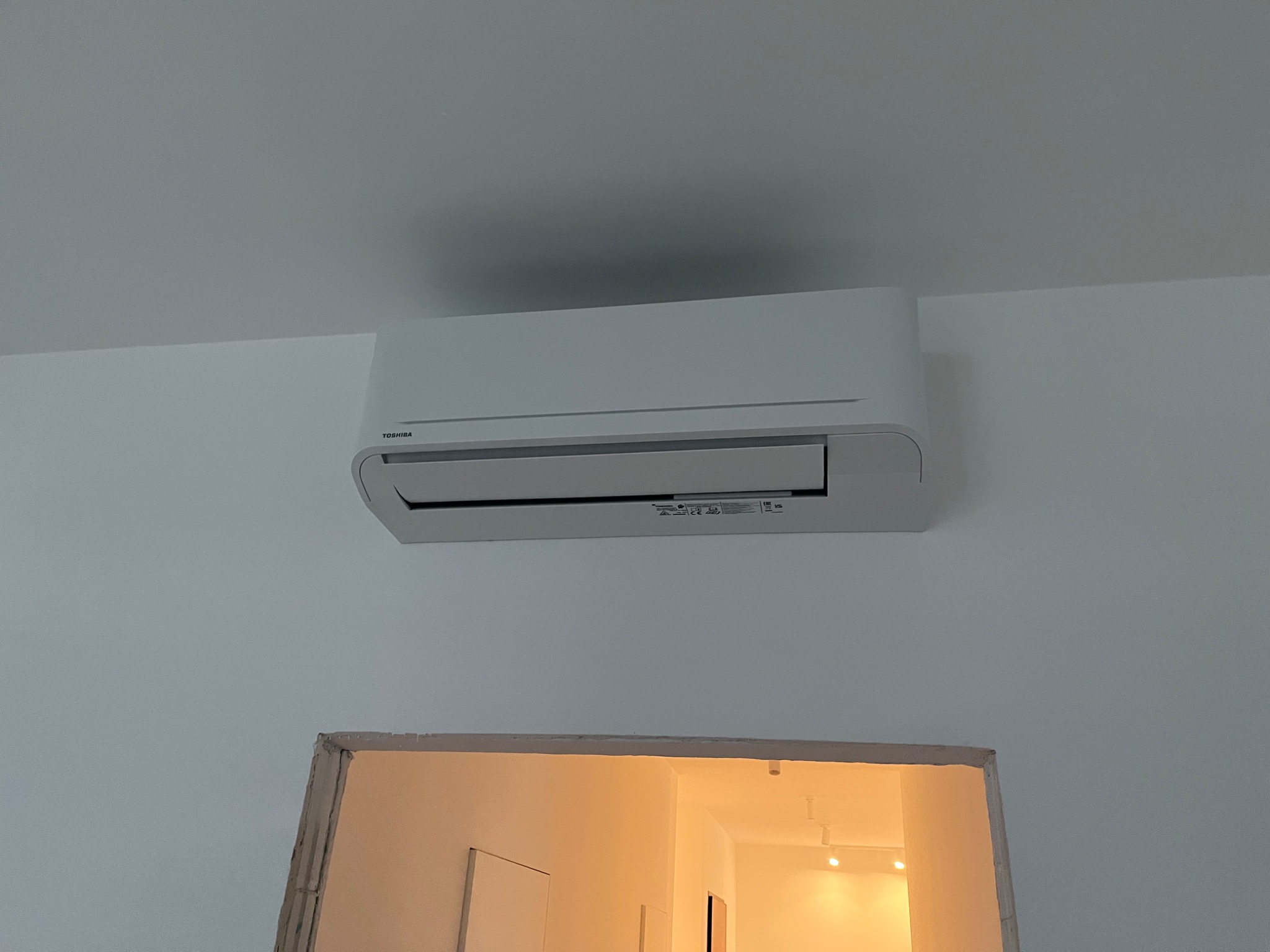 Biała jednostka klimatyzacji Toshiba zamontowana na ścianie nad surowym otworem drzwiowym w jasnym pomieszczeniu.