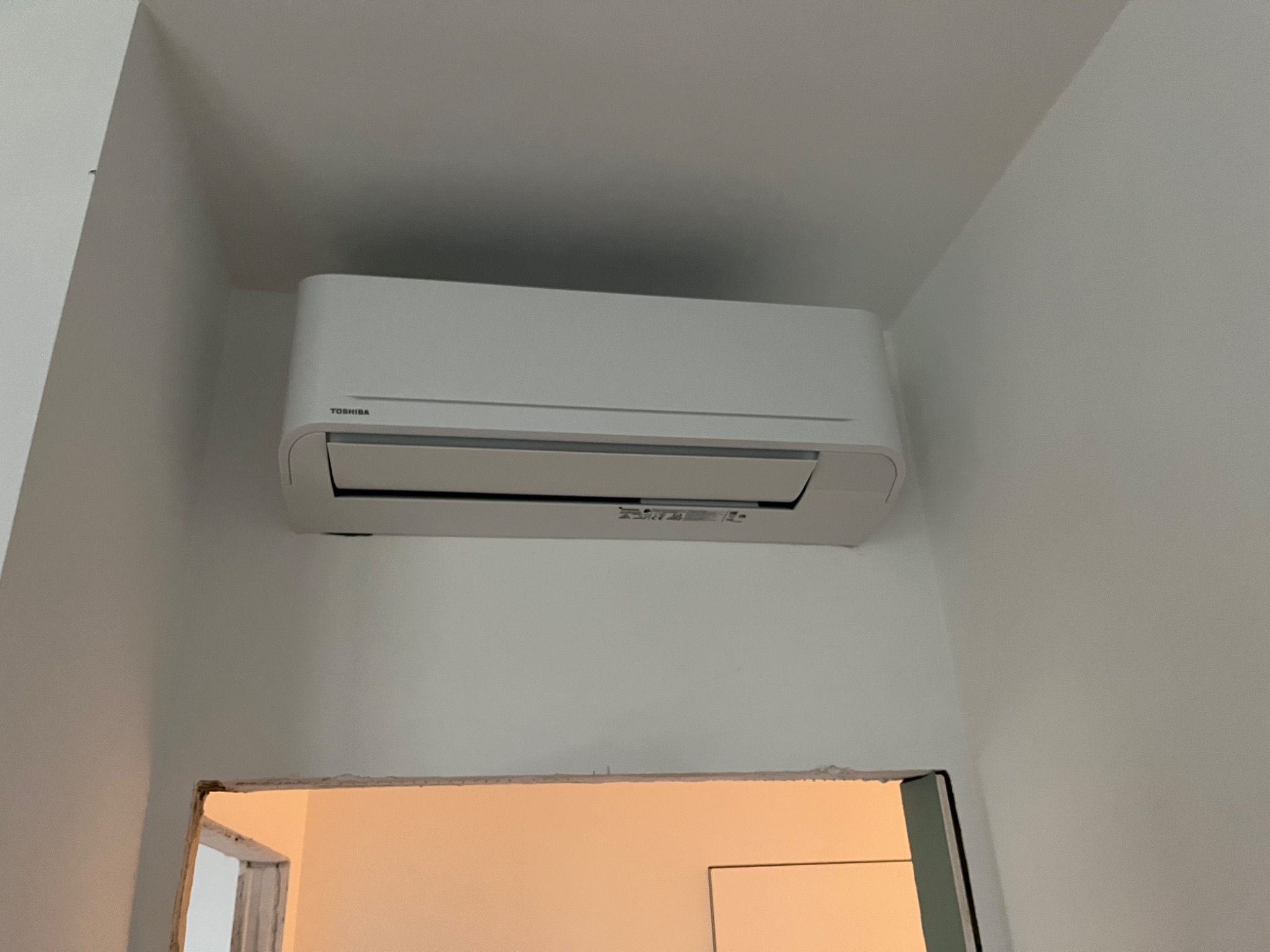 Zamontowana jednostka wewnętrzna klimatyzatora Toshiba na białej ścianie nad surowym otworem drzwiowym, widoczne niedokończone wykończenie.