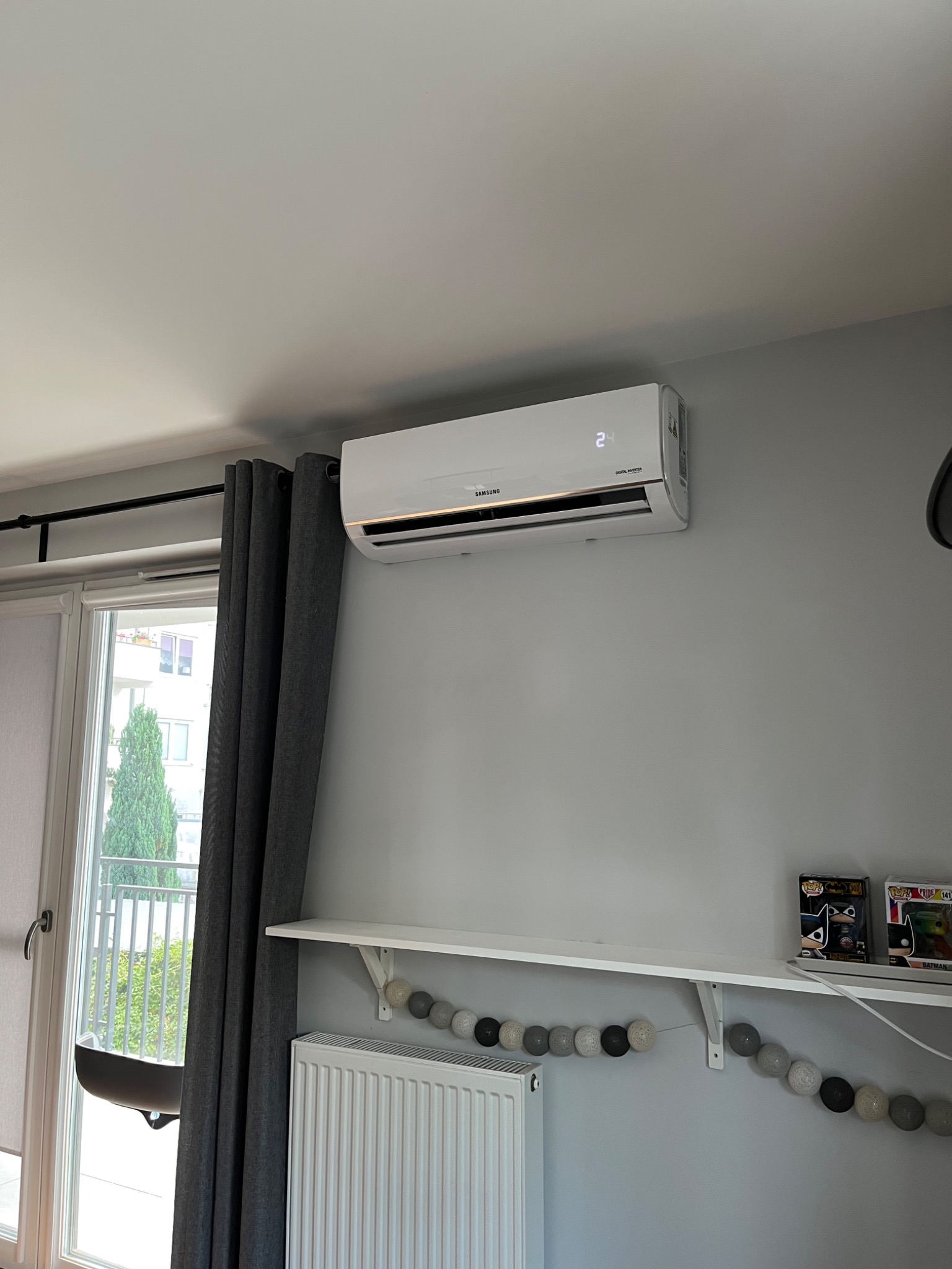 Biała klimatyzacja Samsung Digital Inverter zamontowana na szarej ścianie w pokoju z grzejnikiem, półką z dekoracjami i oknem z widokiem na zieleń.