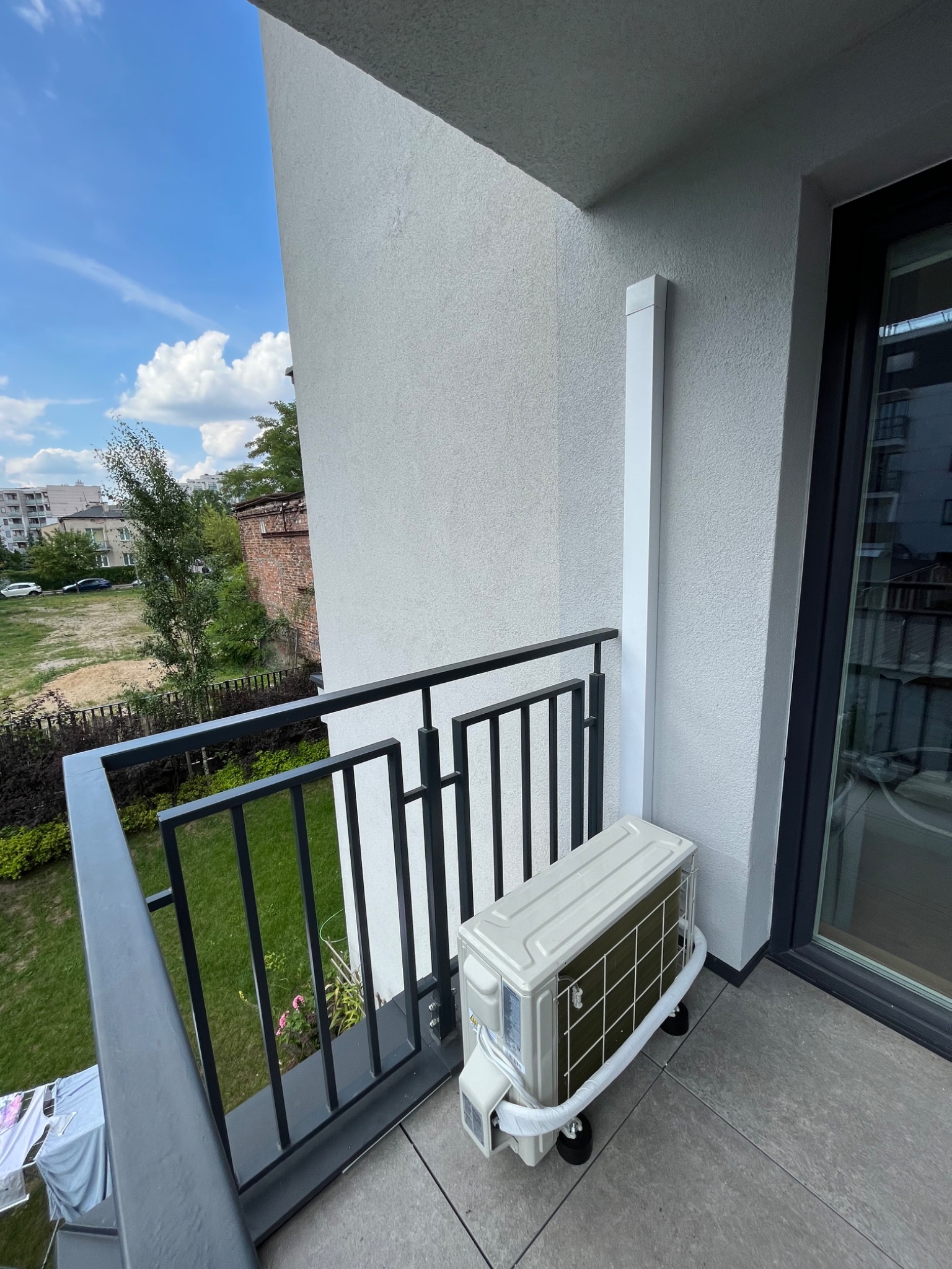 Jednostka zewnętrzna klimatyzatora zamontowana na balkonie mieszkania w bloku, z widocznymi przewodami i elementami montażowymi.