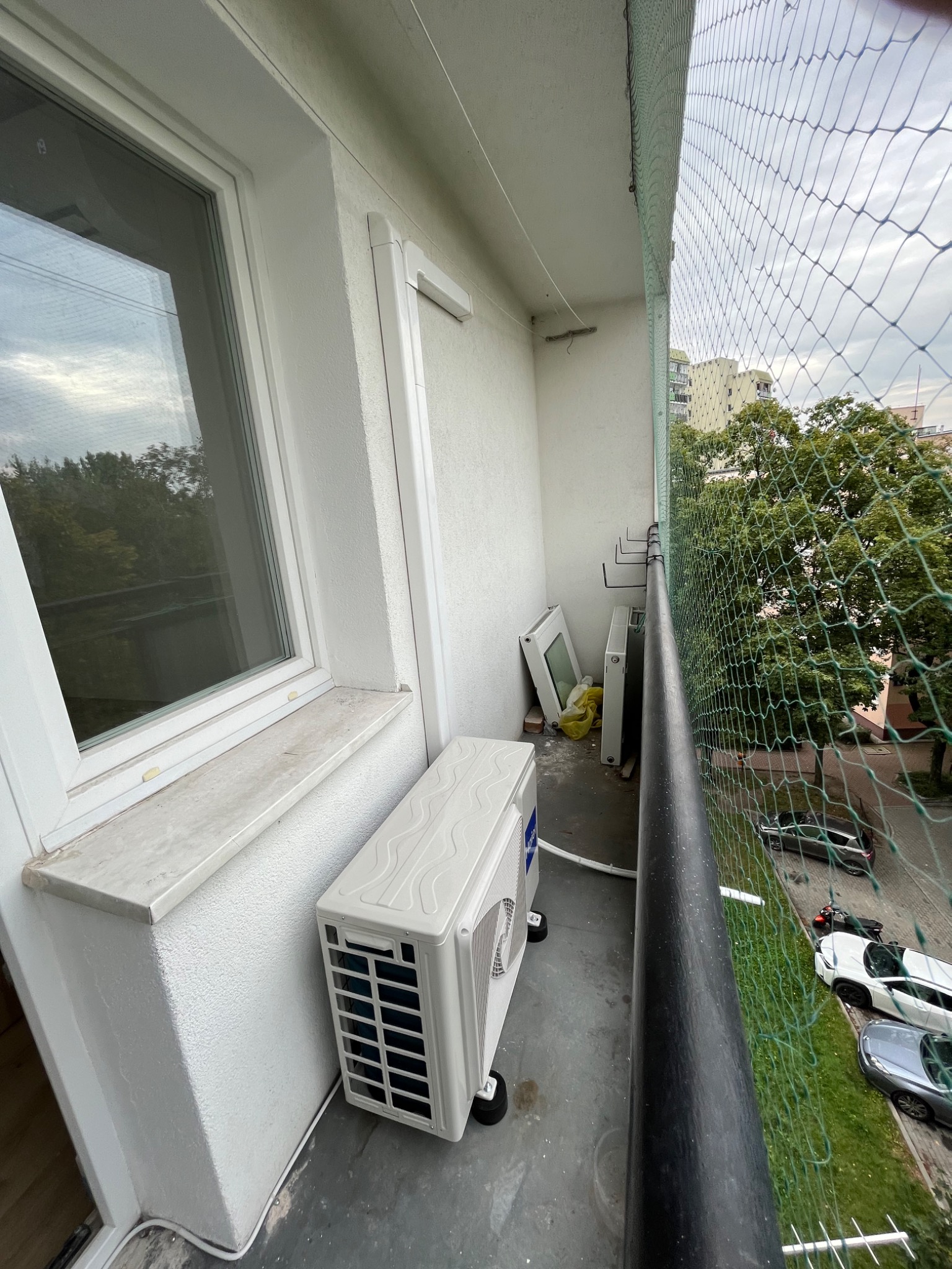 Kompaktowa jednostka zewnętrzna klimatyzatora na balkonie z widokiem na zieleń i parking, zabezpieczonym siatką ochronną.