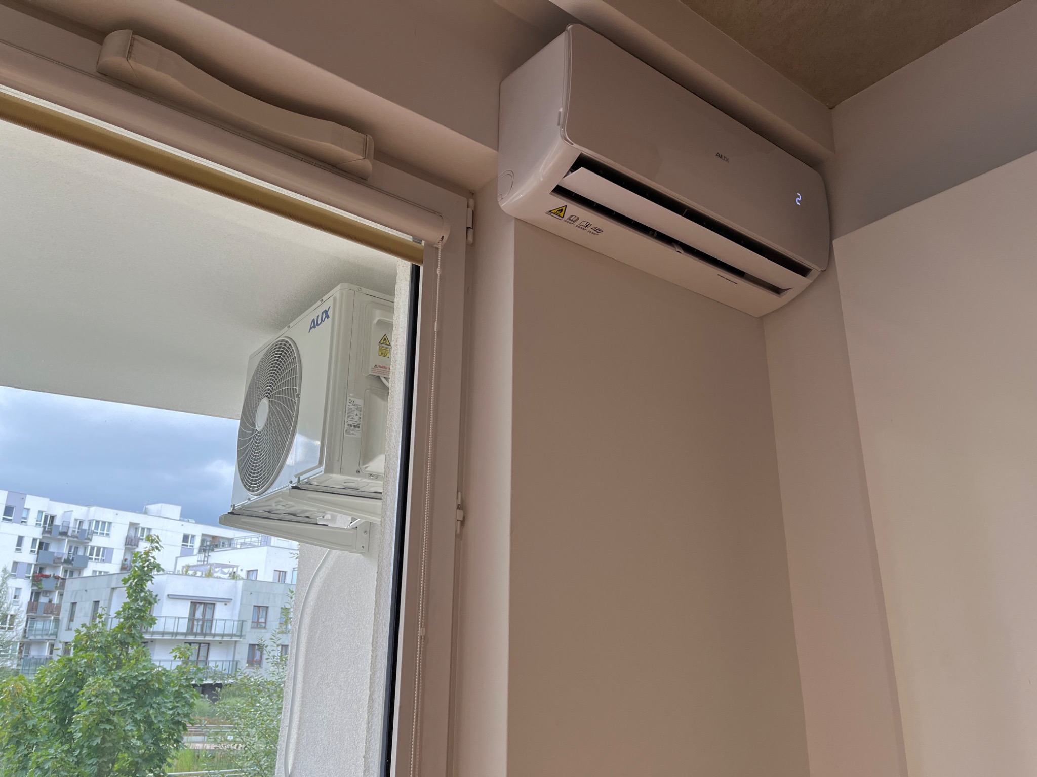 Biała jednostka wewnętrzna klimatyzacji marki AUX zamontowana na ścianie w jasnym pomieszczeniu, widok przez okno na jednostkę zewnętrzną, budynki mieszkalne i zieleń.