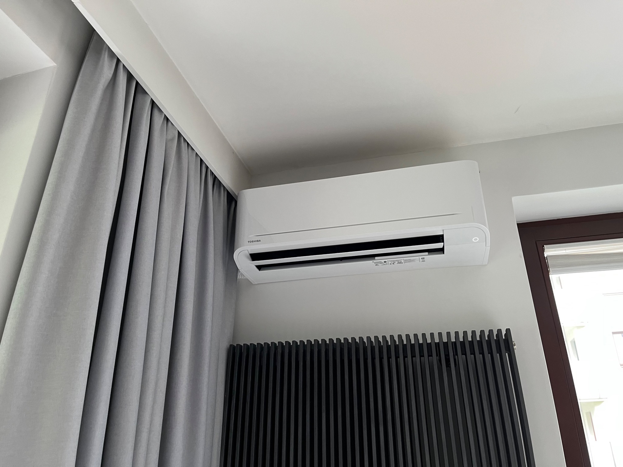 Biała klimatyzacja Toshiba zamontowana na ścianie nad czarnym, designerskim grzejnikiem panelowym, obok okna z roletą i szarych zasłon.