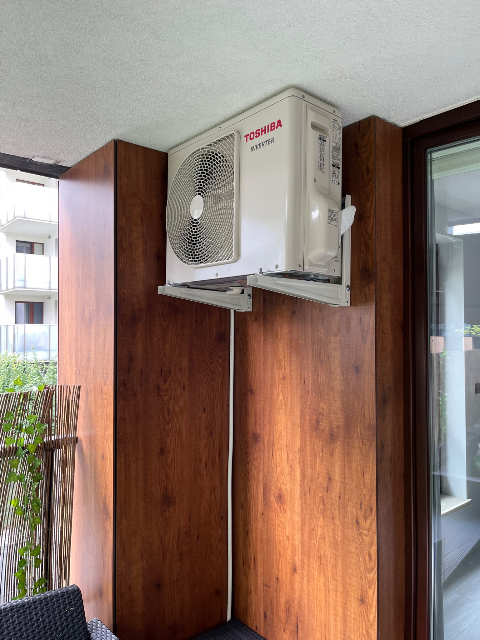 Biała jednostka zewnętrzna klimatyzacji Toshiba Inverter zamontowana na drewnianej obudowie balkonu w mieszkaniu.