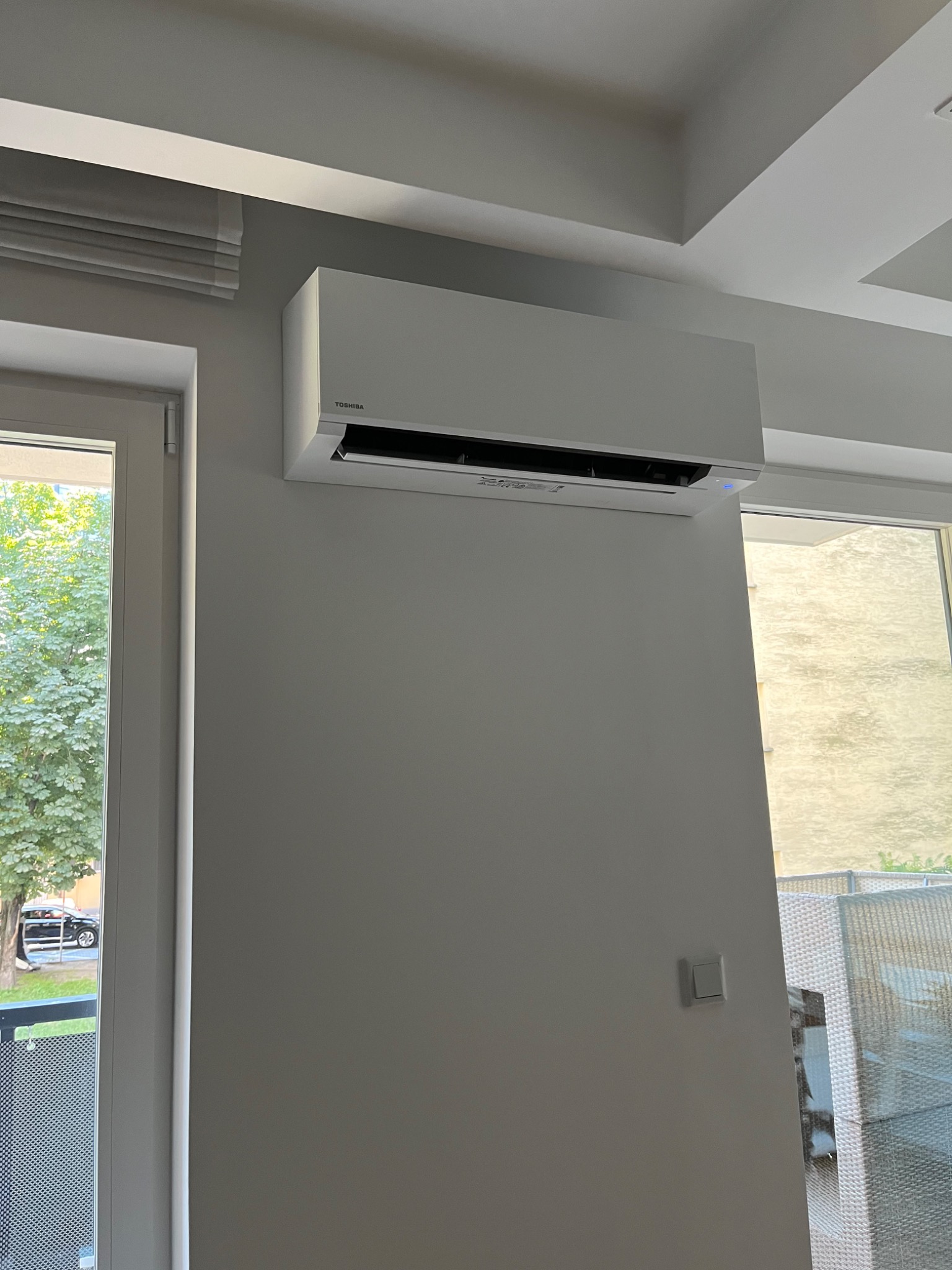 Biała klimatyzacja Toshiba zamontowana na ścianie w jasnym pomieszczeniu mieszkalnym, widoczne okna z roletami i widokiem na zewnątrz.