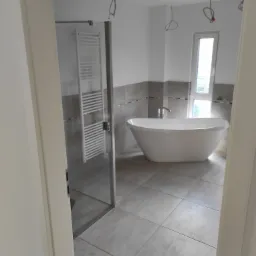 Nowoczesna łazienka z wolnostojącą wanną, grzejnikiem drabinkowym i szarą glazurą, widok przez futrynę drzwiową. Widoczne wystające kable elektryczne.