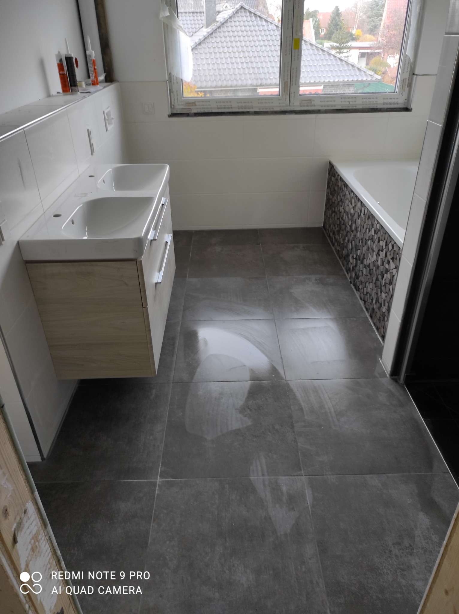 Wykończona łazienka z szarą, błyszczącą podłogą z płytek, umywalką z drewnianą szafką wiszącą i wanną z mozaiką na obudowie.