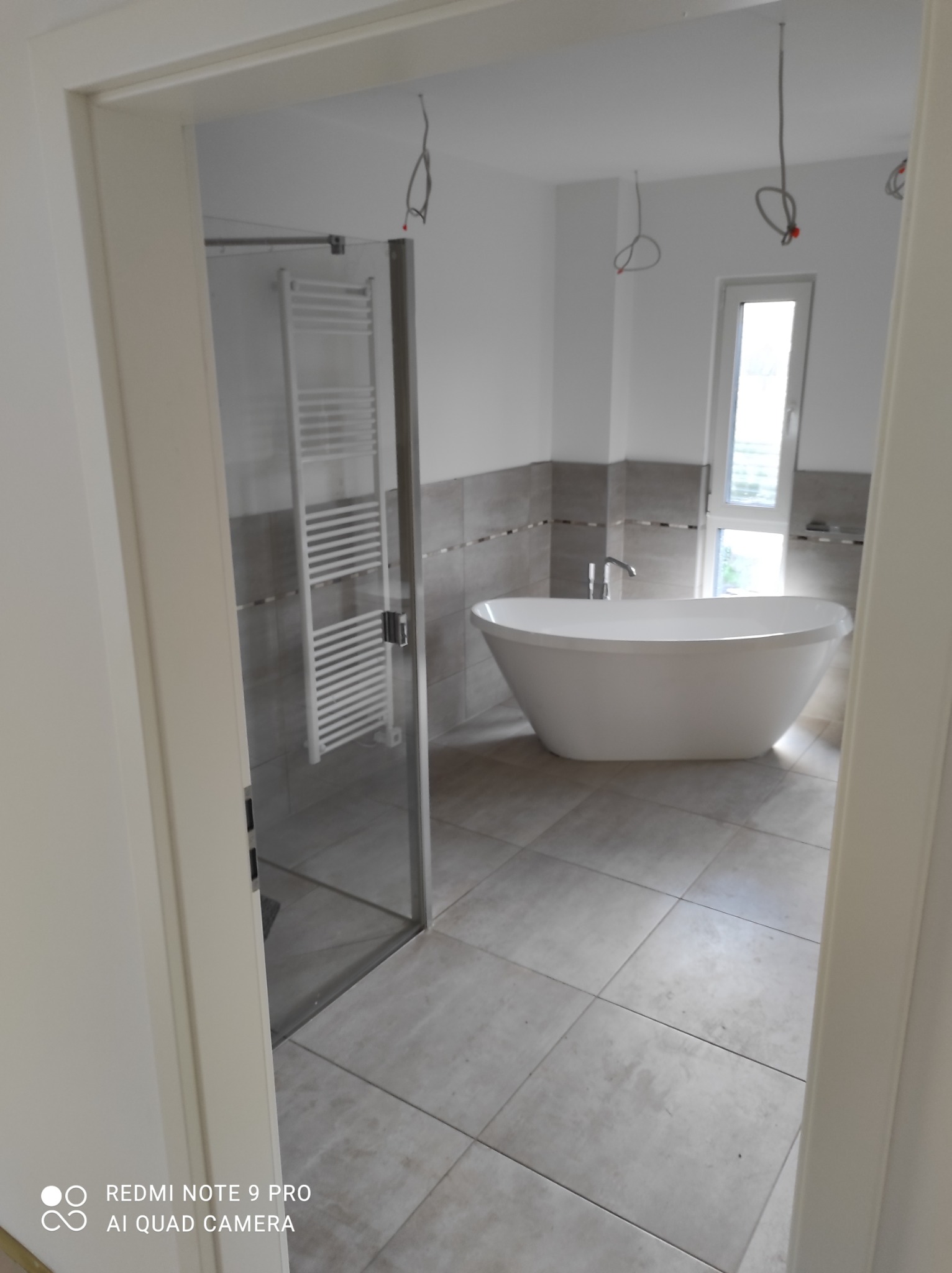 Nowoczesna łazienka z wolnostojącą wanną, grzejnikiem drabinkowym i szarą glazurą, widok przez futrynę drzwiową. Widoczne wystające kable elektryczne.