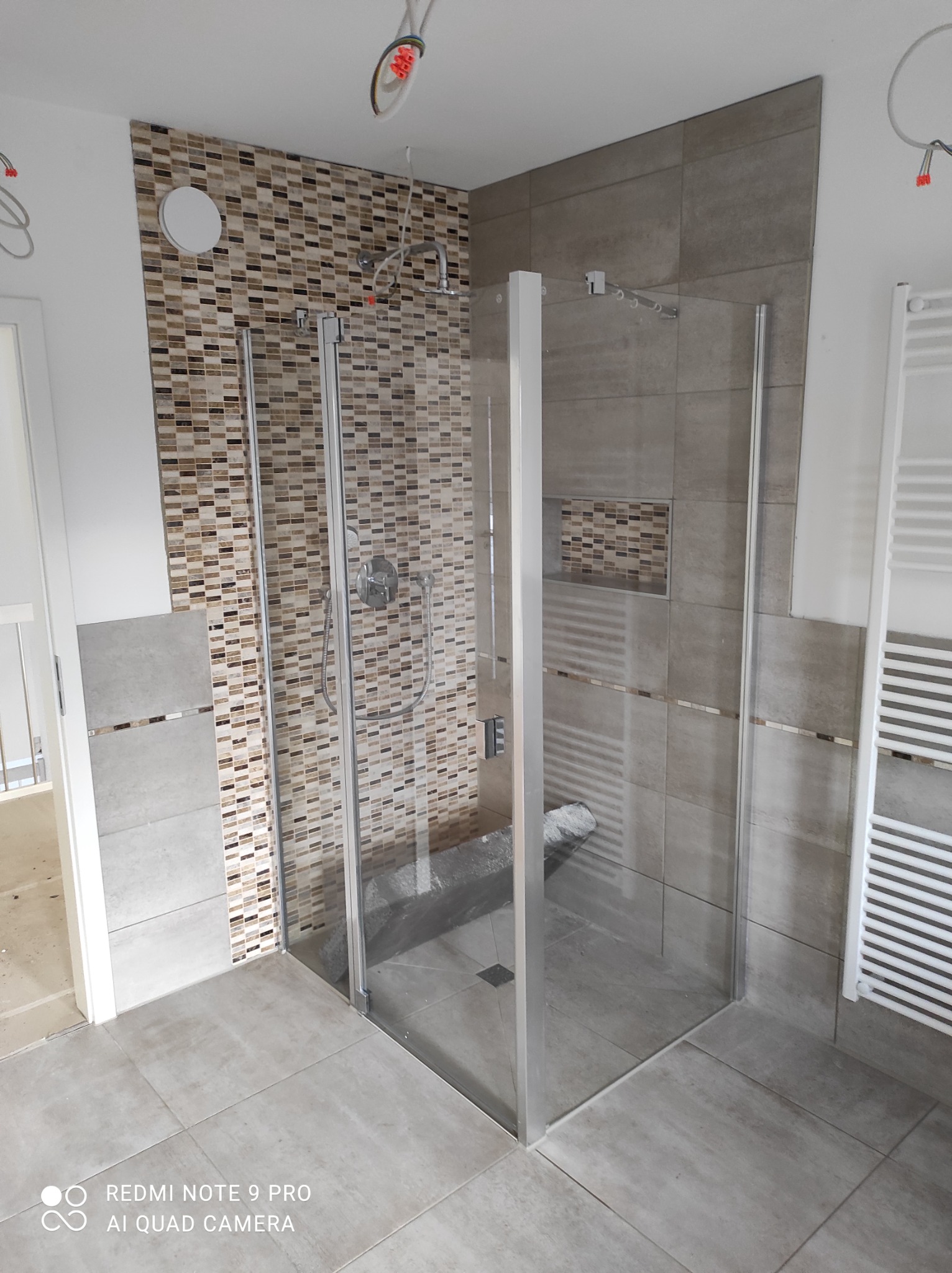 Nowoczesna kabina prysznicowa z mozaiką i szarymi płytkami, widoczne niedokończone prace elektryczne na suficie.