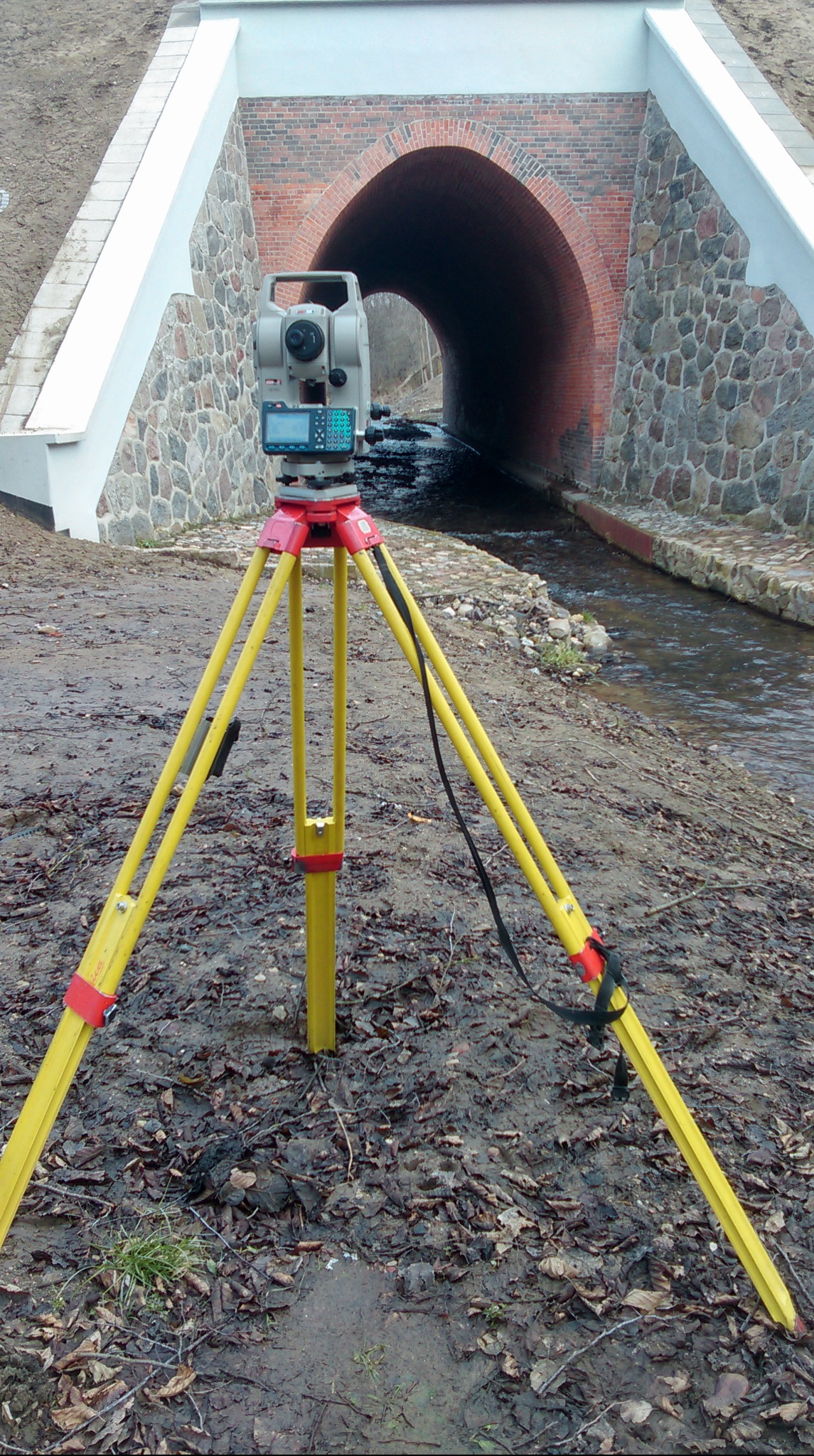 Żółty teodolit na trójnogu ustawiony na błotnistym terenie, z widokiem na ceglany tunel nad strumieniem w tle.