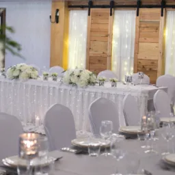 Elegancko udekorowany stół weselny z białymi kwiatami, srebrnymi świecznikami i zastawą, tło z drewnianymi drzwiami stodołowymi i białymi zasłonami z lampkami.