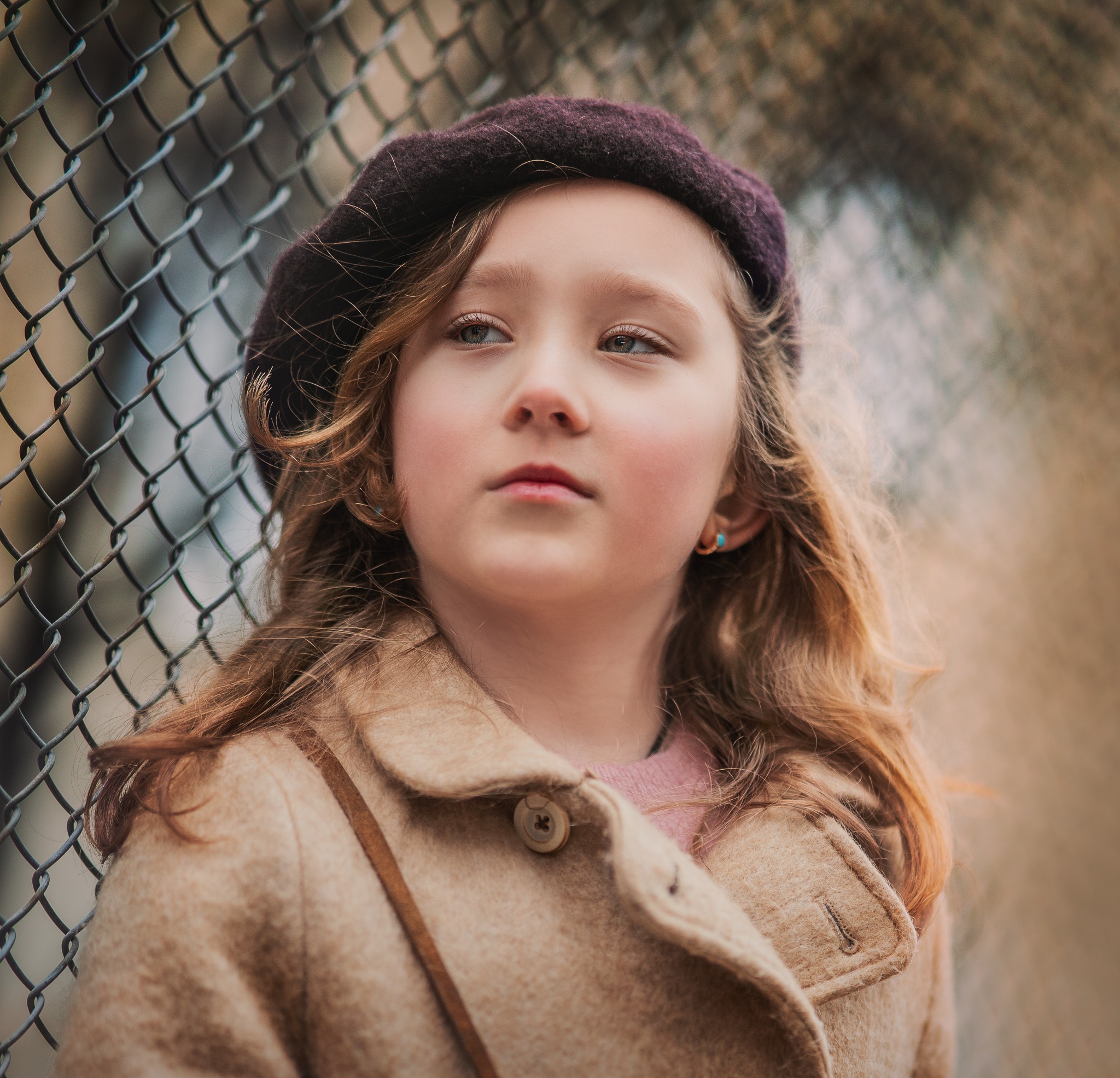 Portret dziewczynki w brązowym berecie i płaszczu, opierającej się o metalową siatkę, zamyślone spojrzenie w górę.
