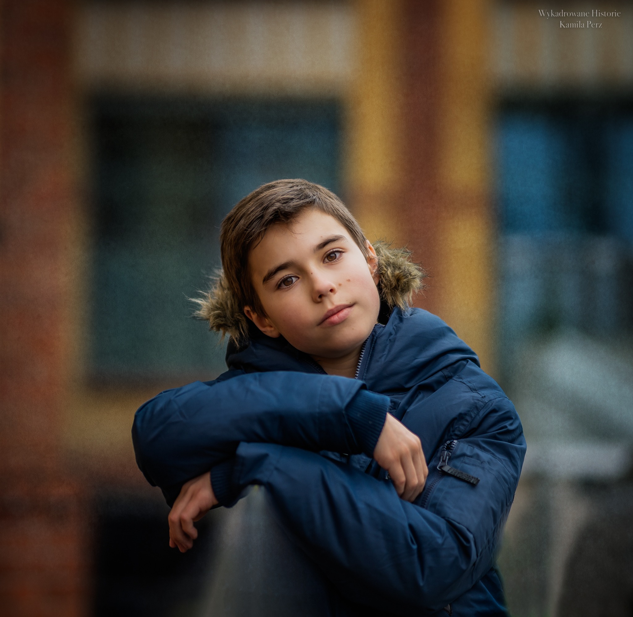Portret młodego chłopca w zimowej kurtce z futrzanym kapturem, opierającego się na szarej powierzchni, z rozmytym, abstrakcyjnym tłem w odcieniach brązu i niebieskiego.