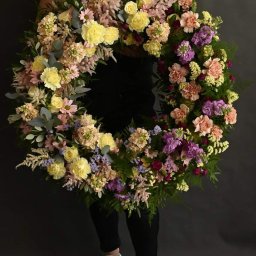 Masz babo kwiatek Izabela Kowalska - Osoba trzyma wieniec z różnokolorowych kwiatów, w tym żółtych róż, fioletowych goździków i zielonych liści, na ciemnoszarym tle. Widoczne białe buty sportowe.