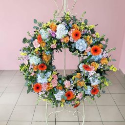 Masz babo kwiatek Izabela Kowalska - Wielobarwny wieniec z gerber, róż, hortensji i eukaliptusa na metalowym stojaku, na tle różowej ściany i kremowej podłogi z płytek.