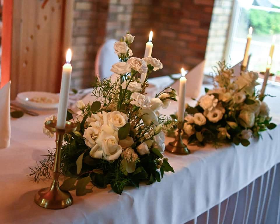 Dekoracja stołu z białych róż i świec w złotych świecznikach, ustawiona na białym obrusie z plisami, w tle ceglana ściana i okno.