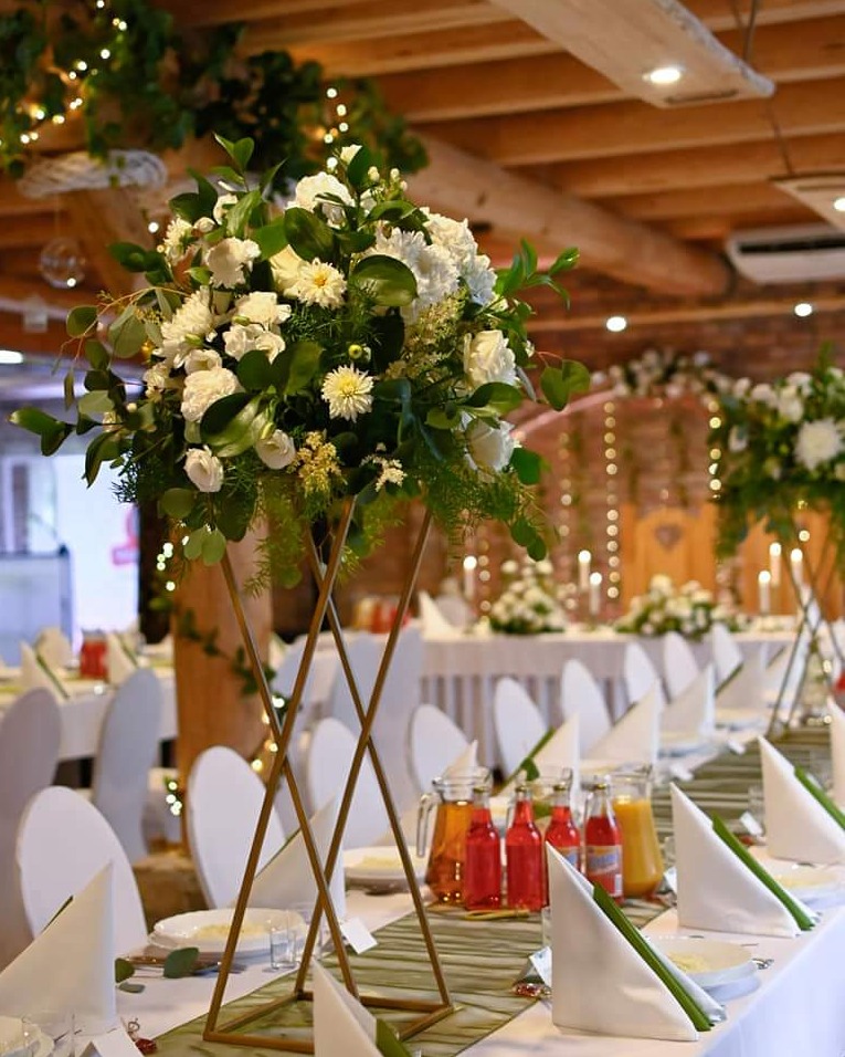 Dekoracja stołu weselnego z wysokim wazonem z białych kwiatów i zielenią, napoje w szklanych naczyniach, białe krzesła i oświetlenie dekoracyjne w tle.