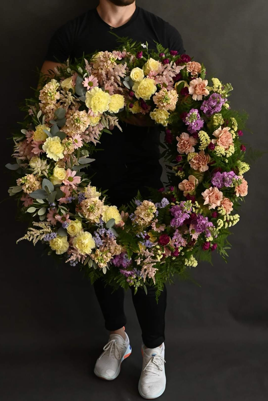 Osoba trzyma wieniec z różnokolorowych kwiatów, w tym żółtych róż, fioletowych goździków i zielonych liści, na ciemnoszarym tle. Widoczne białe buty sportowe.