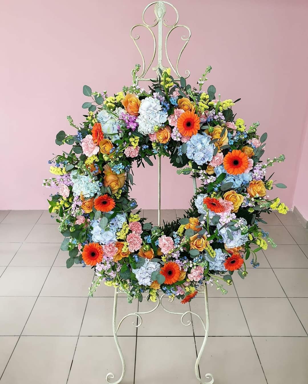 Wielobarwny wieniec z gerber, róż, hortensji i eukaliptusa na metalowym stojaku, na tle różowej ściany i kremowej podłogi z płytek.