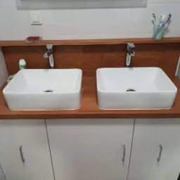 Dwie białe, kwadratowe umywalki nablatowe z chromowanymi bateriami, zamontowane na drewnianym blacie w kolorze orzecha, osadzonym na białej szafce z minimalistycznymi uchwytami w łazience.