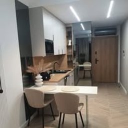 Fabryka Snów Adam Pawlicki - Jasna kuchnia w aneksie, z drewnianym blatem, czarną armaturą i dekoracyjnym wazonem z suszonymi trawami na blacie. Widoczne dwie krzesła barowe i podłoga ułożona w jodełkę.