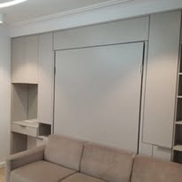 Fabryka Snów Adam Pawlicki - Wielofunkcyjna zabudowa ścienna w odcieniach szarości, z ukrytym łóżkiem, biurkiem i półkami, uzupełniona beżową sofą w dolnej części kadru.