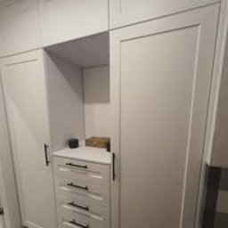 Fabryka Snów Adam Pawlicki - Biała zabudowa meblowa z ciemnymi uchwytami, obejmująca szafę z przesuwnymi drzwiami i komodę z szufladami, uzupełniona otwartą półką z drobnymi przedmiotami.