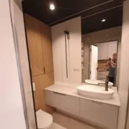 Nowoczesna łazienka z zabudową meblową z jasnego drewna, białą umywalką nablatową o owalnym kształcie i minimalistyczną armaturą w kolorze czarnym; w lustrze odbija się osoba robiąca zdjęcie.