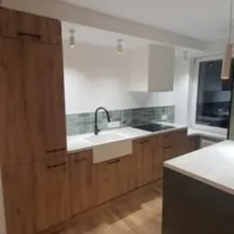 Nowoczesna kuchnia z drewnianymi frontami szafek, białym zlewem farmerskim i szarymi płytkami nad blatem. Widoczne minimalistyczne oświetlenie punktowe i biały okap. Blat jasny, imitujący kamień.