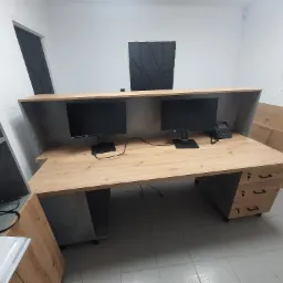 Biurko recepcyjne z blatem w odcieniach drewna, wyposażone w dwa monitory i telefon, z szafką z trzema szufladami na klucz, ustawione w jasnym pomieszczeniu.