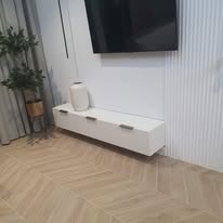 Biała, wisząca szafka RTV z minimalistycznymi uchwytami na tle ściany z pionowymi panelami dekoracyjnymi i parkietu ułożonego w jodełkę francuską. Obok szafki stoi roślina doniczkowa, a nad nią...