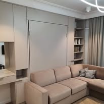 Beżowa sofa narożna przed zabudową meblową z ukrytym łóżkiem, jasne wnętrze z półkami na książki i biurkiem z lustrem.